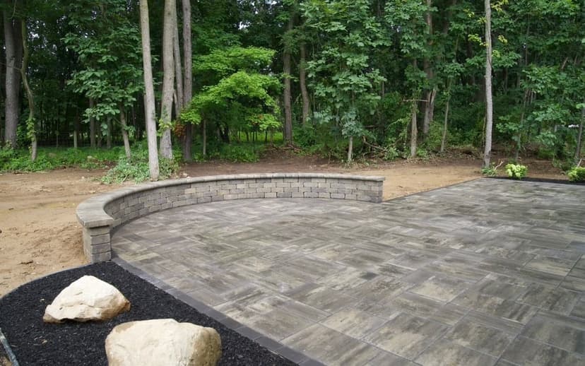 Paved outdoor patio with curved stone wall and landscaping in a forested area.