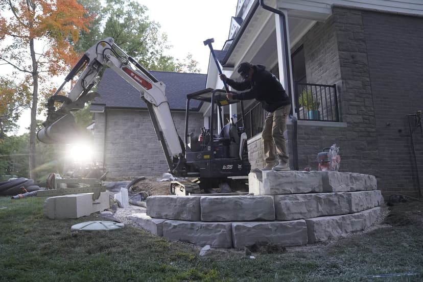 Construction worker operating a mini excavator to build a stone wall in a residential yard.