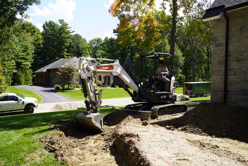 Excavator digging a trench in a residential yard with trees and a house in the background.