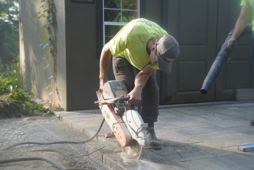 Worker using a concrete saw on a stone patio, wearing a safety helmet and neon shirt.