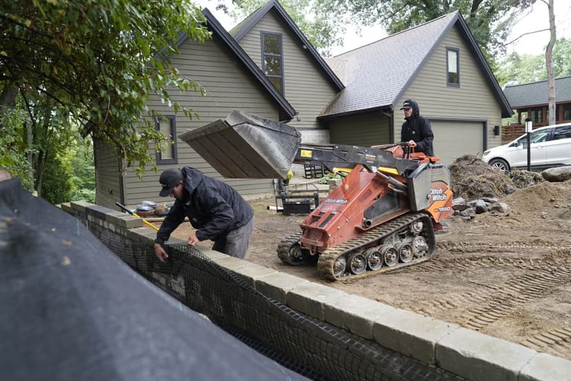 Construction workers using machinery to build a stone wall beside a house.