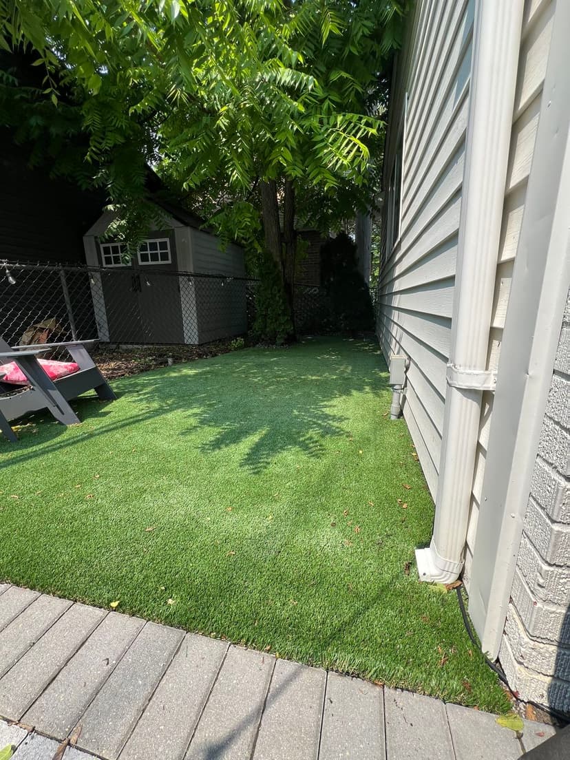 Artificial grass area beside a house, surrounded by trees and a stone pathway.