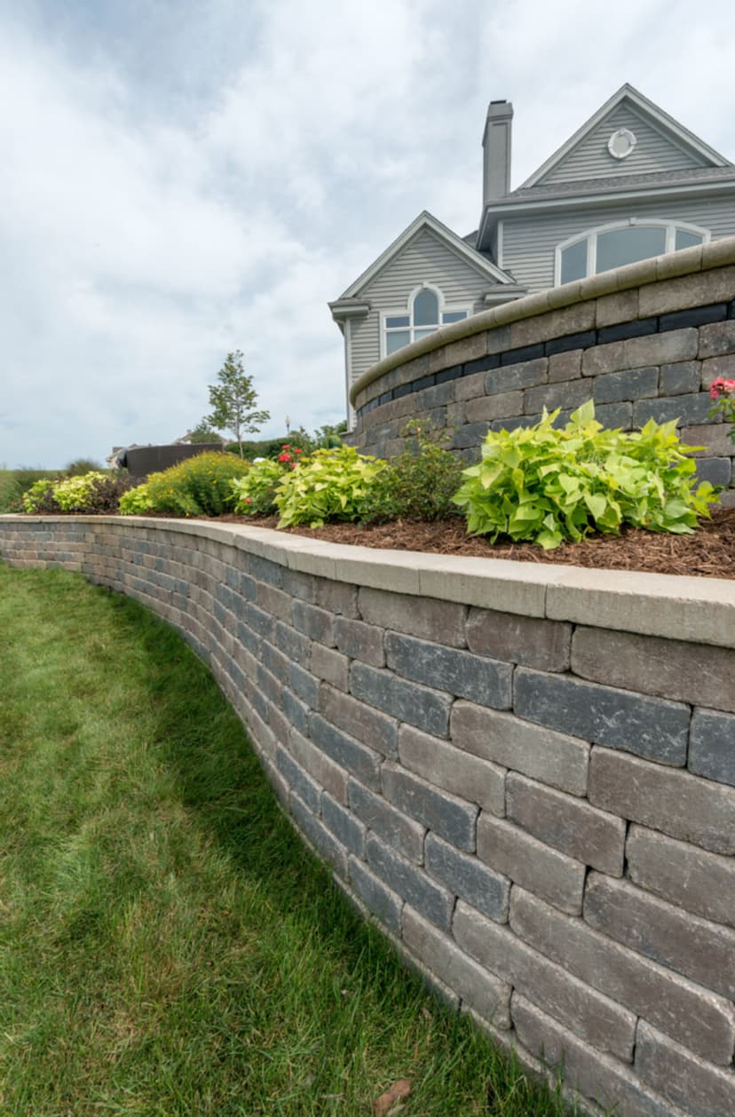 Curved stone retaining wall with colorful flowers and a gray house in the background.