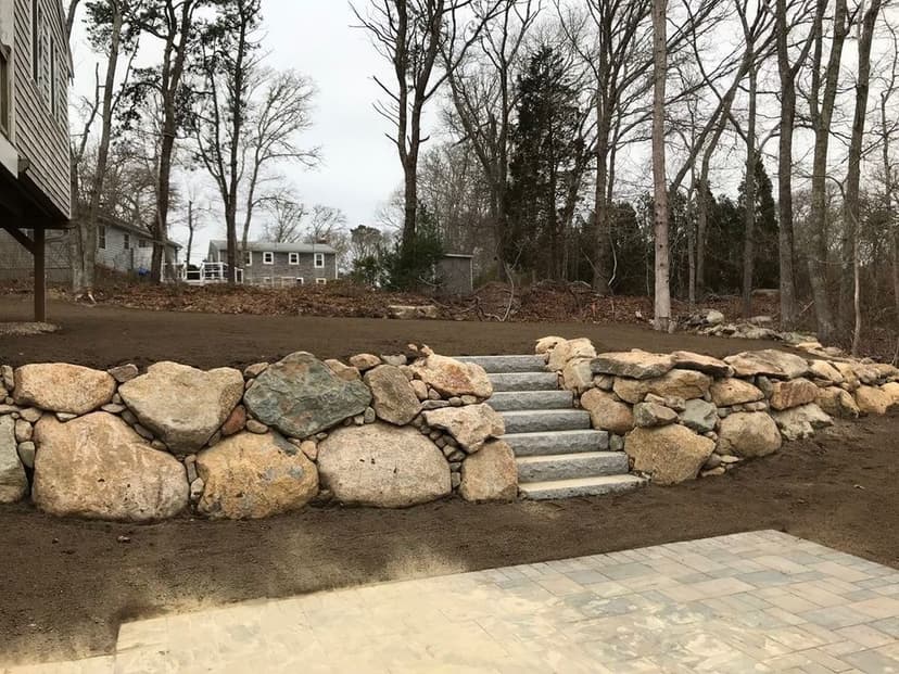 Stone landscaping with steps leading up from a patio to a wooded area in a residential backyard.