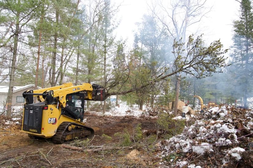 Excavator clearing trees in a forested area with smoke and snow visible.