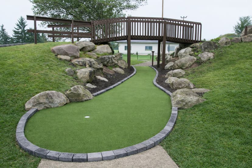 Mini golf course pathway leading to a wooden bridge surrounded by rocks and lush grass.