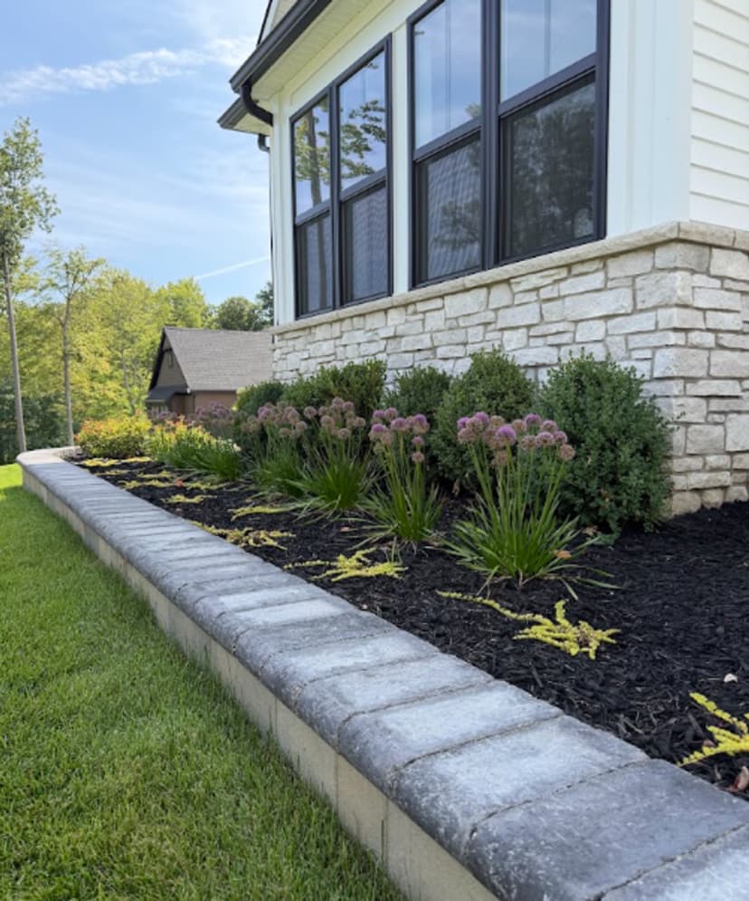 Stone garden border with vibrant flowers and greenery beside a house exterior.