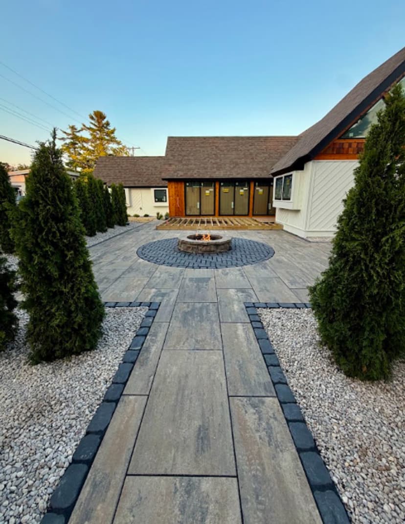 Modern patio with fire pit, bordered by greenery and elegant stone pathway, leading to a stylish house.