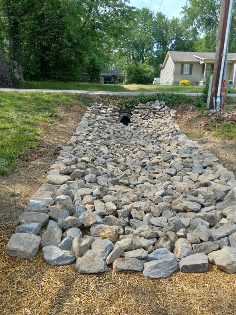 Stone drainage channel lined with rocks, leading to a culvert in a residential area.